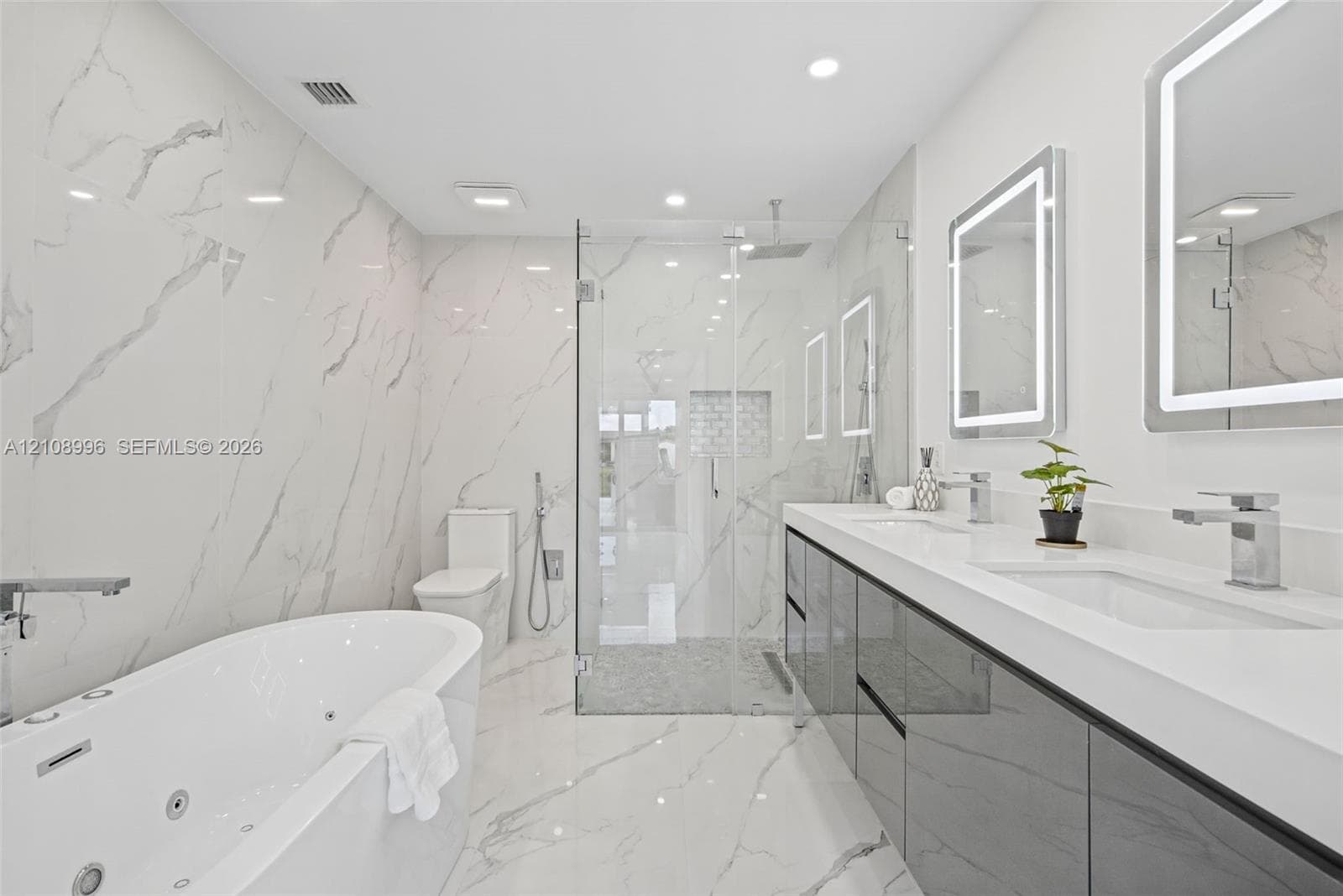 Full-wall veined stone-look tiling, jetted soaking tub, dual-sink vanity with high-gloss cabinetry, glass-enclosed walk-in shower with stone tile flooring, and integrated LED-lighted mirrors