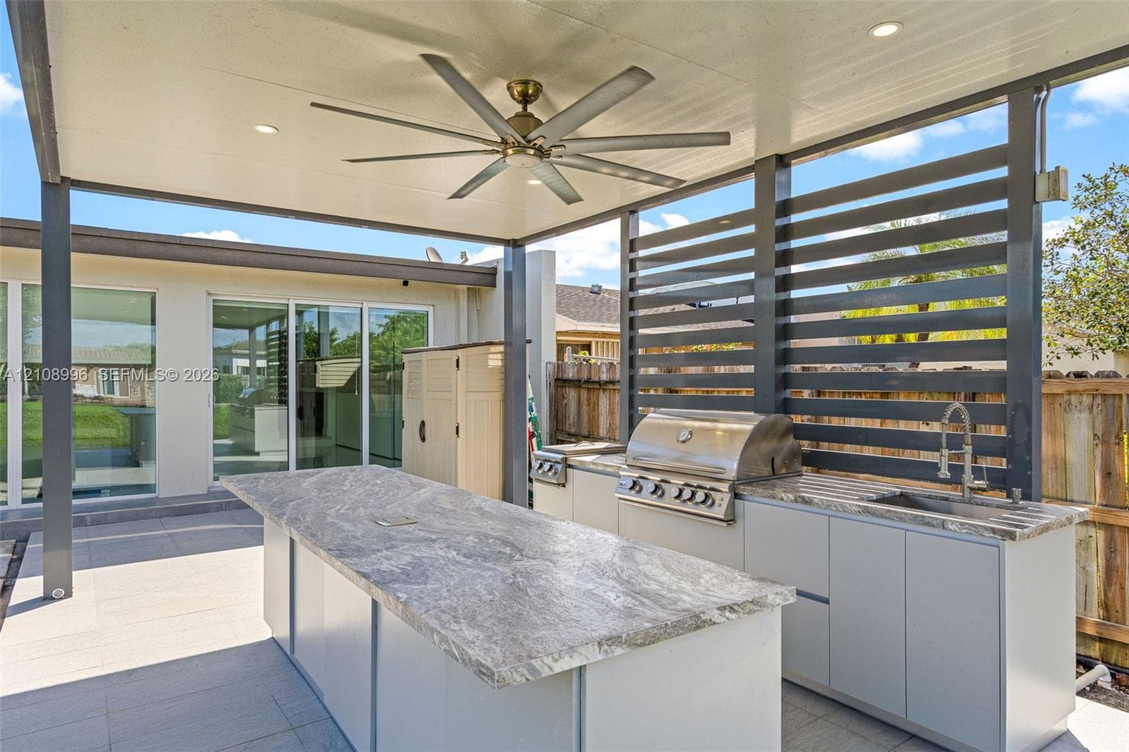 Covered outdoor kitchen featuring stone-top counters, a stainless steel built-in grill, a sink, and a multi-blade ceiling fan under a structured patio roof