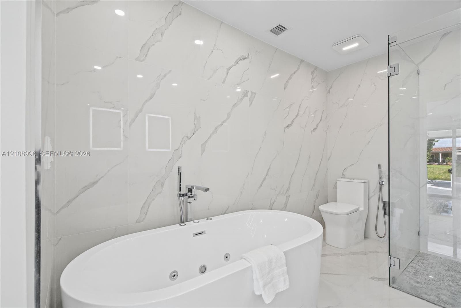 Freestanding jetted soaking tub with deck-mounted chrome faucet, floor-to-ceiling large-format stone-finish tile wall, frameless glass enclosed shower, and white dual-flush toilet