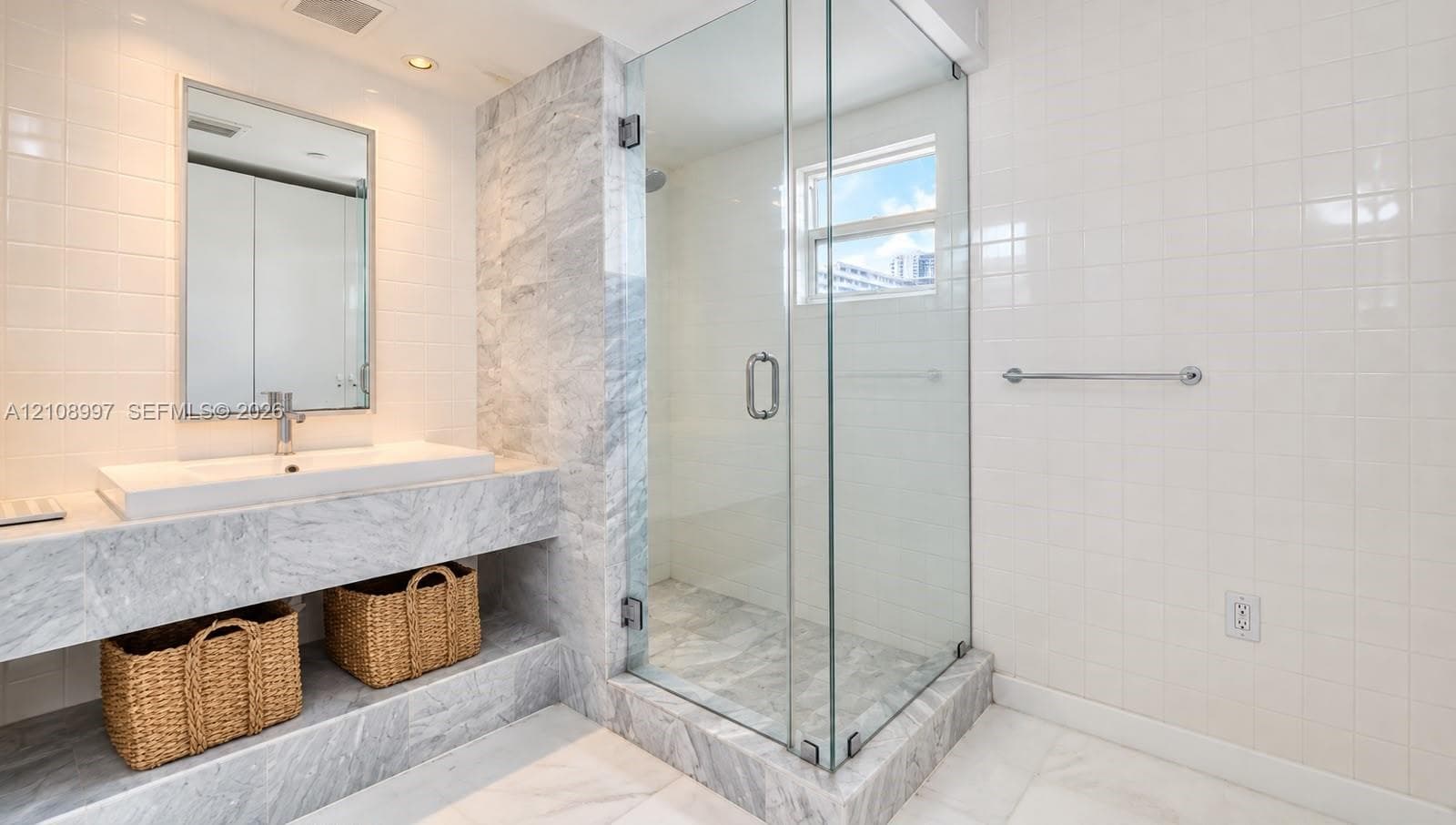 Stone-surfaced vanity with an integrated rectangular sink, framed wall mirror, glass-enclosed shower, and neutral tile wall treatments