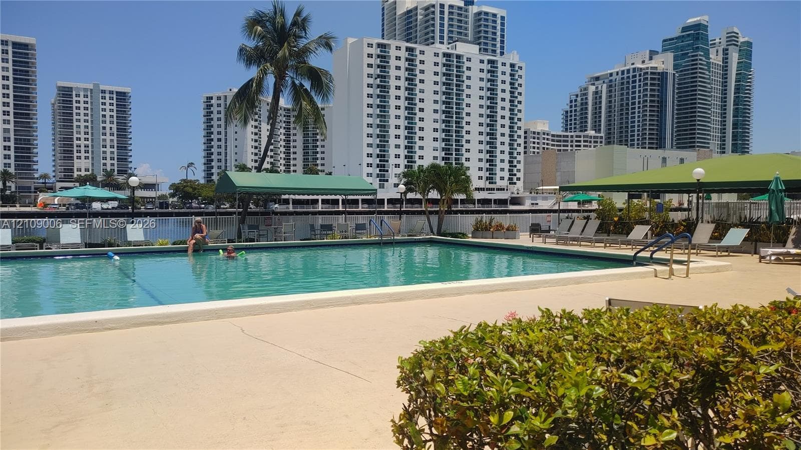 In-ground swimming pool featuring a concrete deck, perimeter fencing, overhead canopy structures, and mature landscaping
