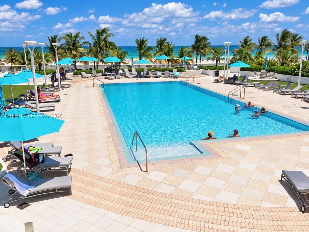 Rectangular swimming pool featuring a tiled deck, built-in steps, and metal handrails, situated against an ocean backdrop with mature palm trees
