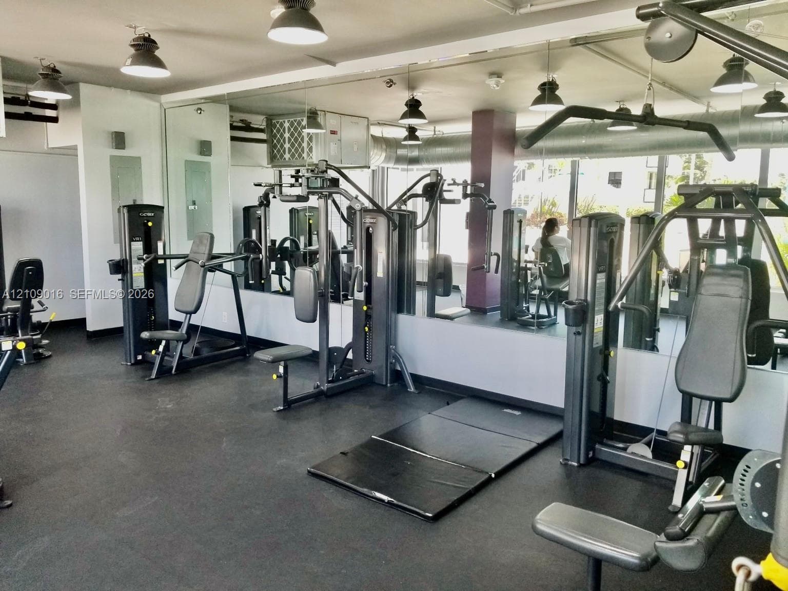 Fitness center featuring rubberized flooring, expansive wall-to-wall mirrors, industrial-style exposed ceiling ductwork, and overhead pendant lighting