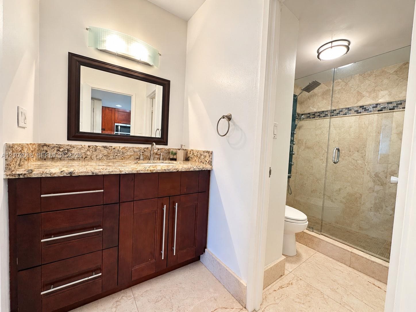 Bathroom featuring dark wood-finish cabinetry, stone countertop, glass-enclosed shower with decorative tile inlay, and stone flooring