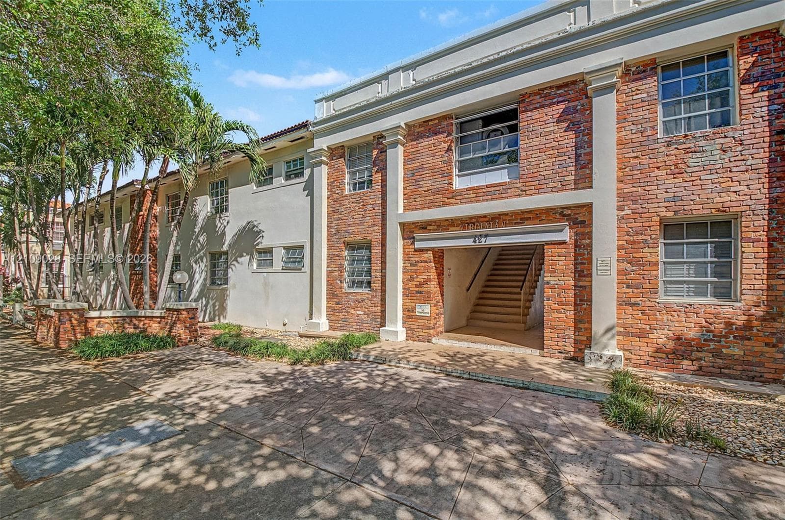 Multi-story exterior featuring a red brick facade, decorative columns, multi-pane windows, and a paved concrete entrance path