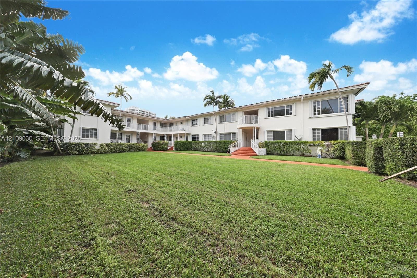 Multi-story building exterior featuring a white stucco facade, expansive manicured lawn, terracotta-toned entry stairs, and mature tropical landscaping