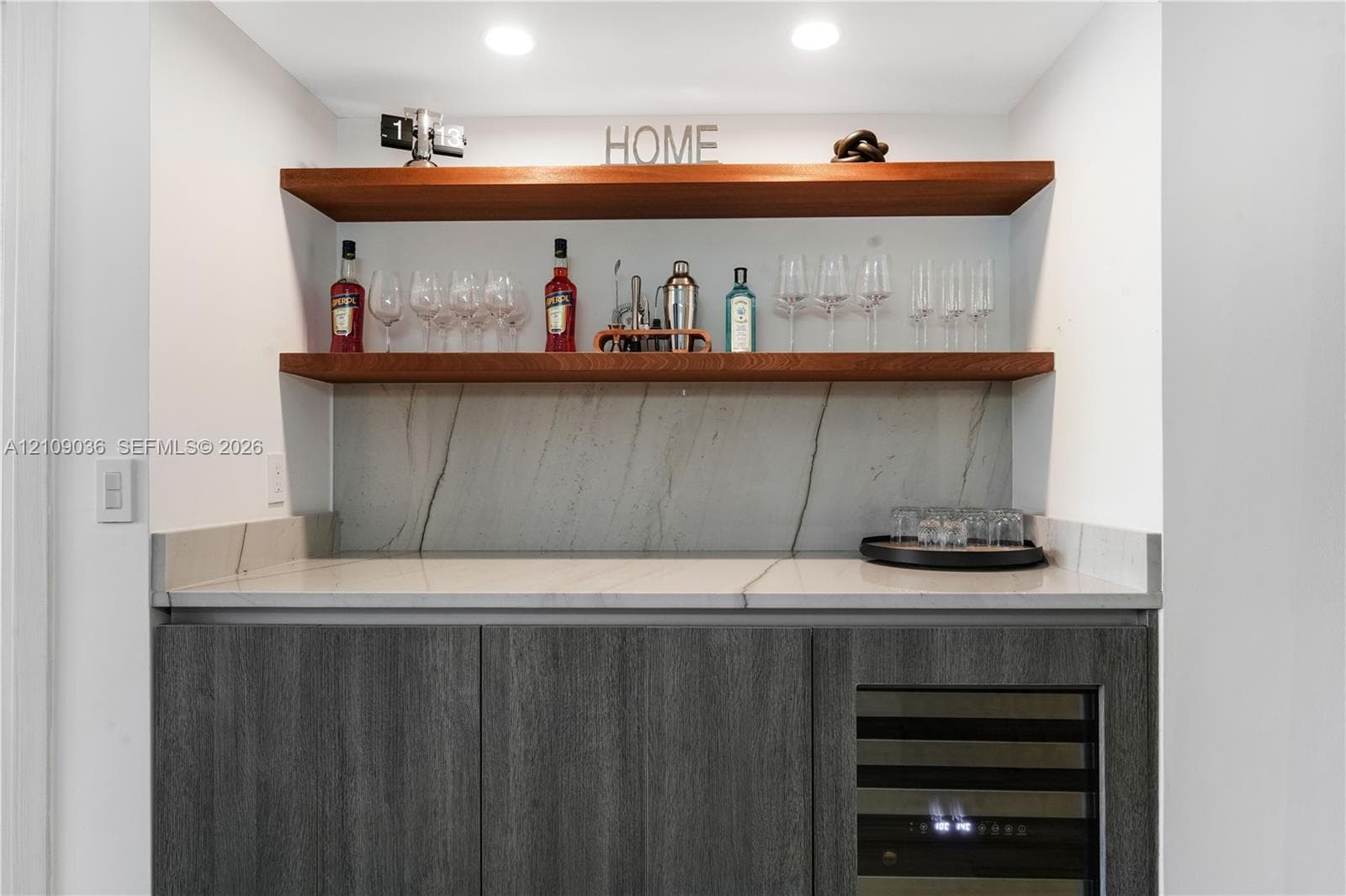 Custom wet bar featuring charcoal-toned cabinetry, a stone-patterned countertop and backsplash, dual wood-finish floating shelves, and a built-in wine refrigerator