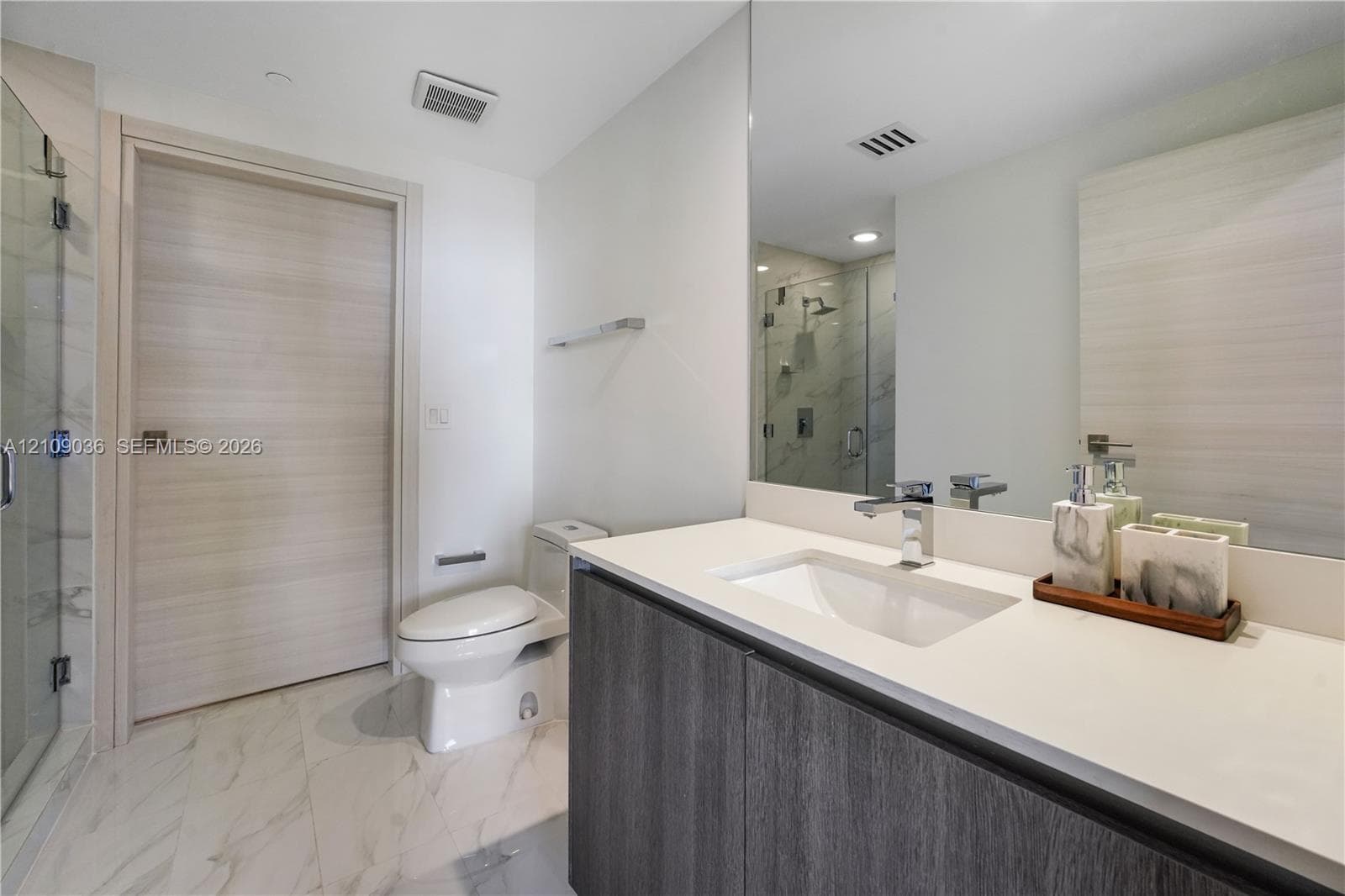 Modern vanity with a light-toned countertop, integrated basin, and dark wood-finish cabinetry, paired with large-format stone-finish floor tile, a full-width mirror, and a glass-enclosed shower