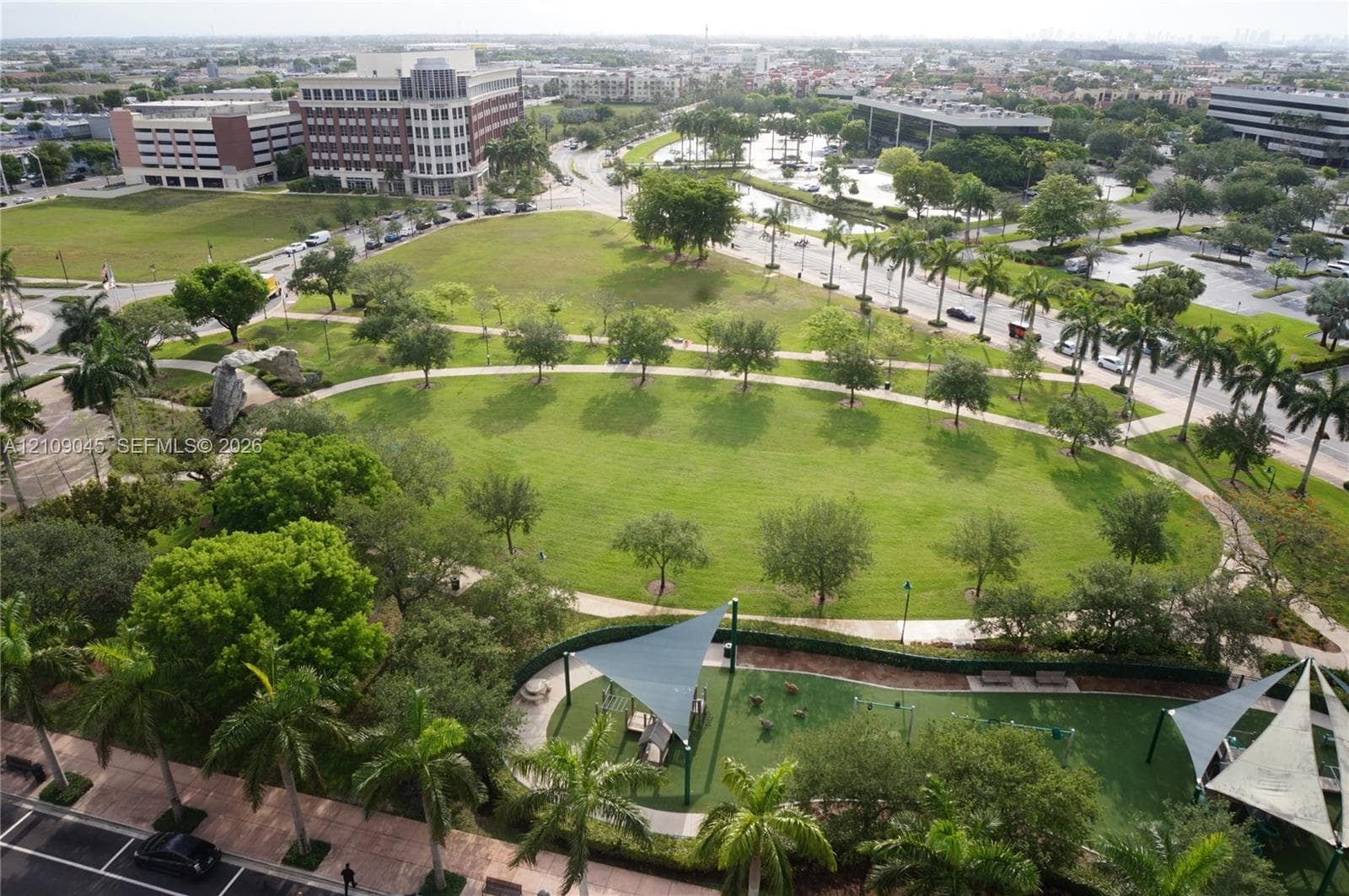 Expansive green space featuring mature trees, paved walking paths, curated landscaping, and shaded recreation structures