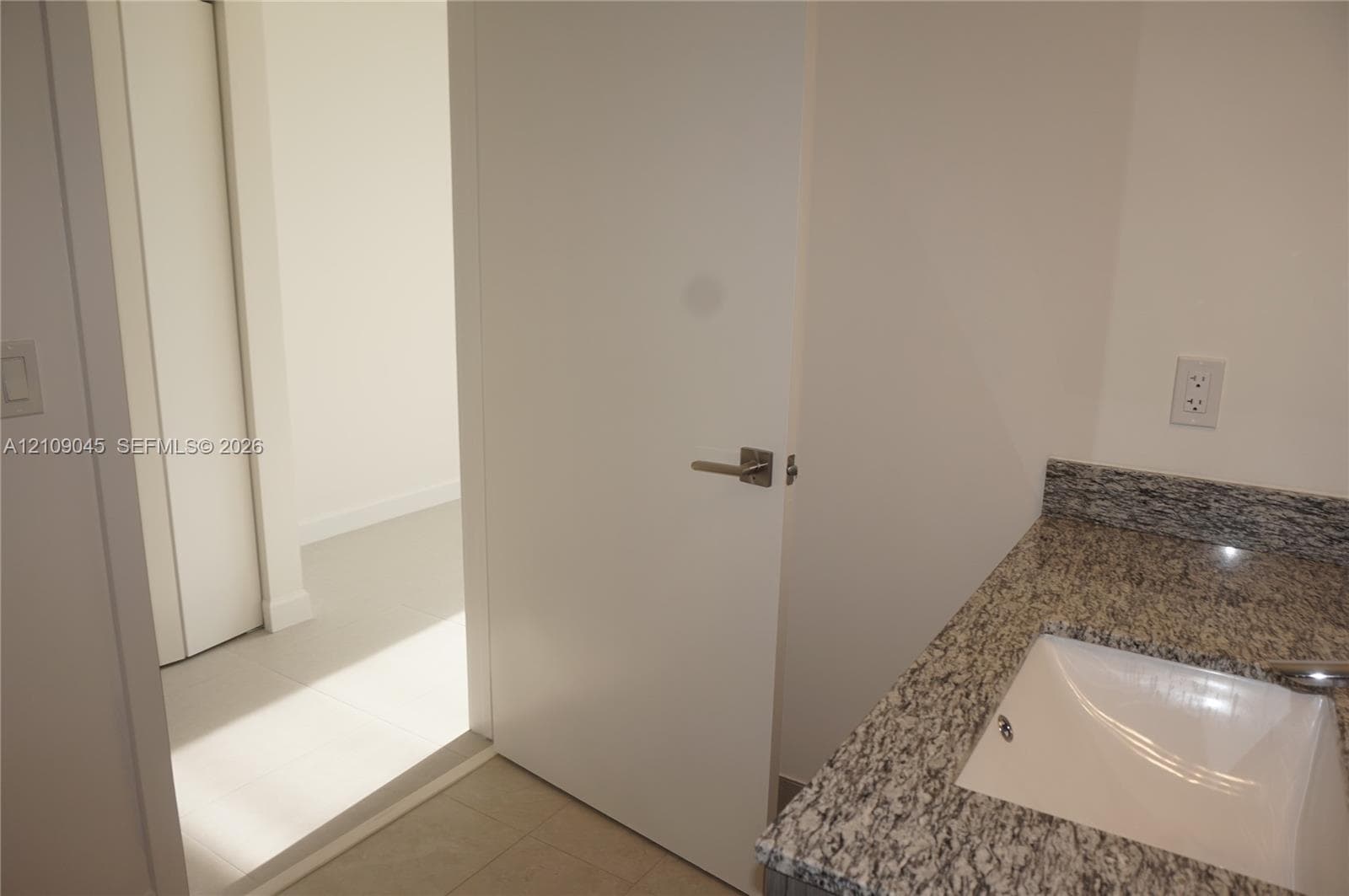 Stone-finish countertop featuring an integrated rectangular sink and modern metal hardware, complemented by neutral tile flooring and a solid-core door with a matte metallic lever handle