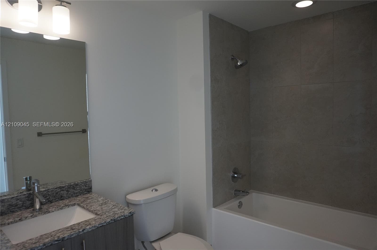 Bathroom featuring a vanity with a stone-finish countertop and integrated sink, chrome faucet, large frameless mirror, and a tub with a tile-surround wall