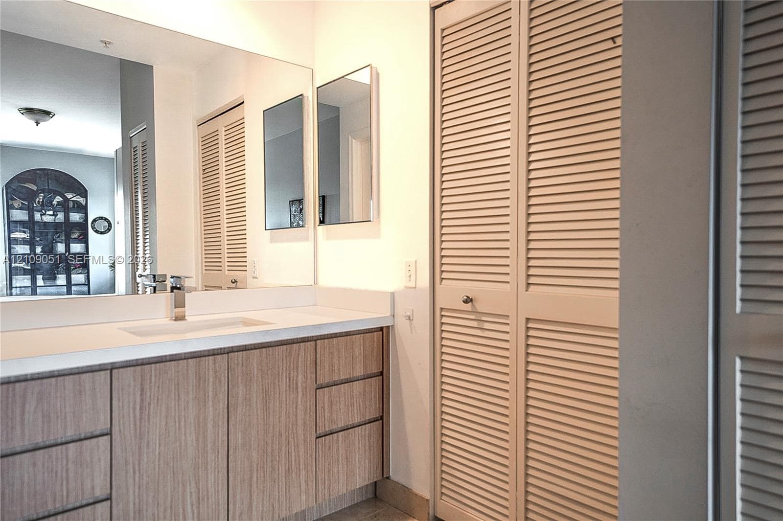 Vanity with wood-finish cabinetry, white countertop, integrated rectangular sink, wall-spanning mirror, and bi-fold louvered closet doors