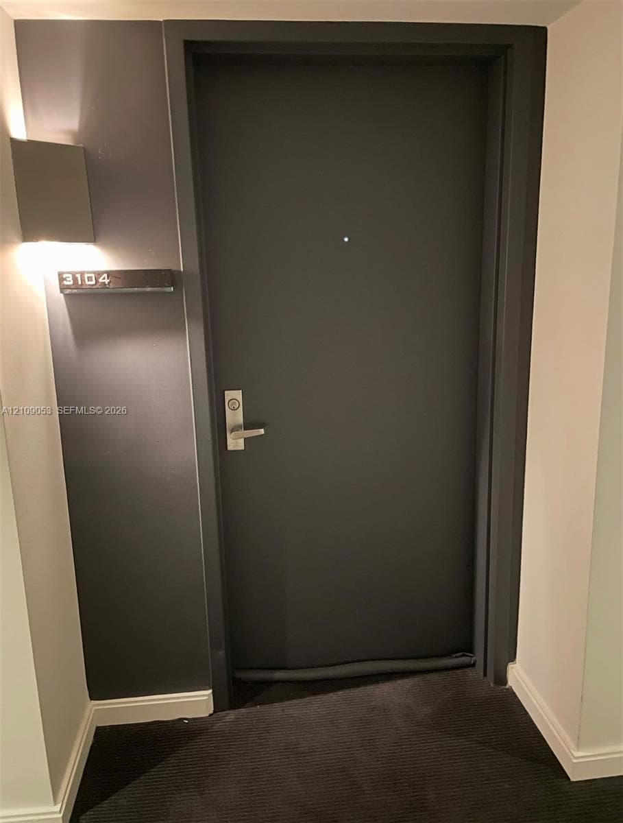 Solid entry door with a metallic handle set and a peephole, featuring a rectangular wall-mounted light fixture and a modern address plaque