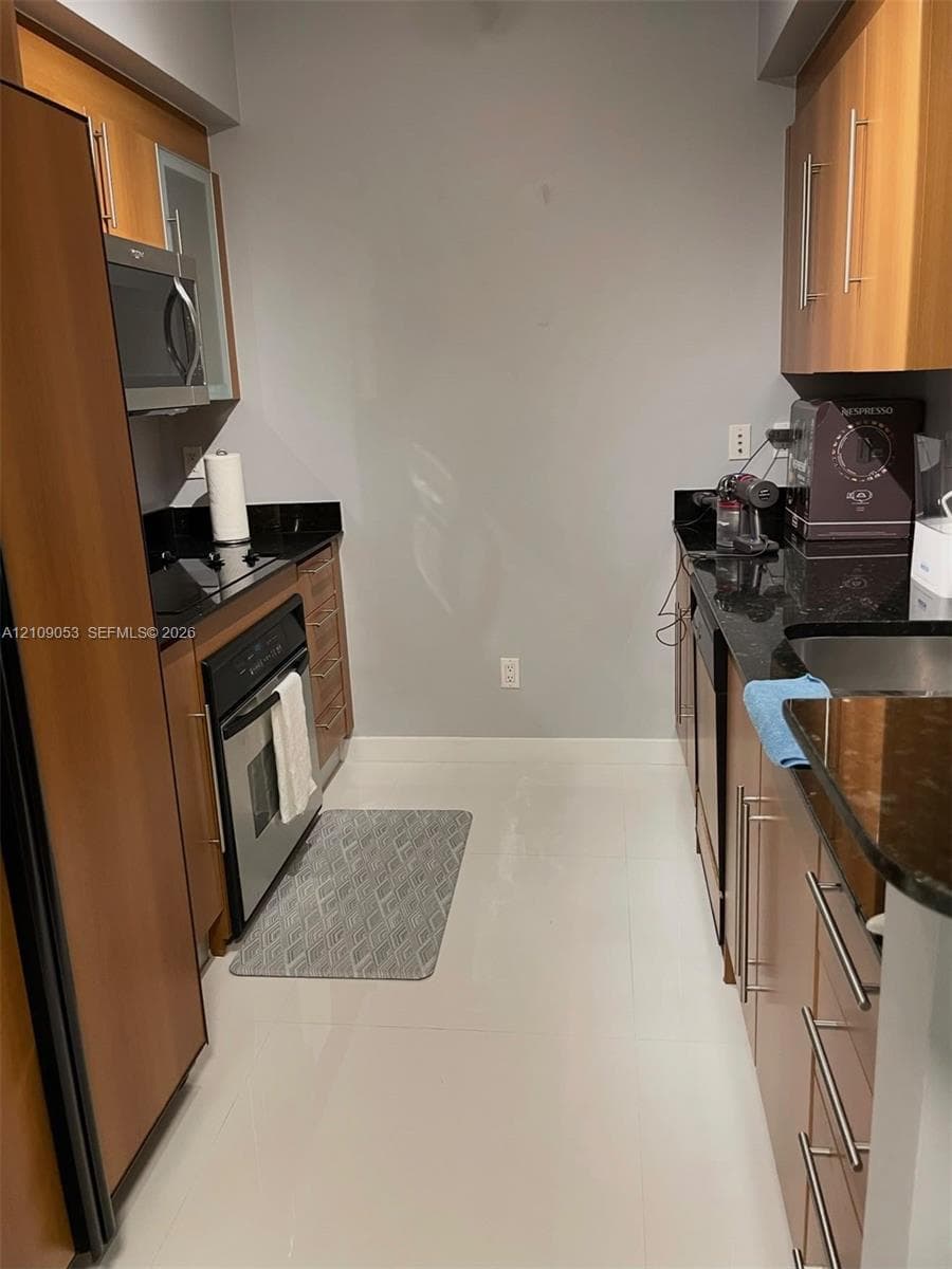 Galley kitchen featuring wood-finish cabinetry, black stone countertops, stainless steel appliances, and tile flooring