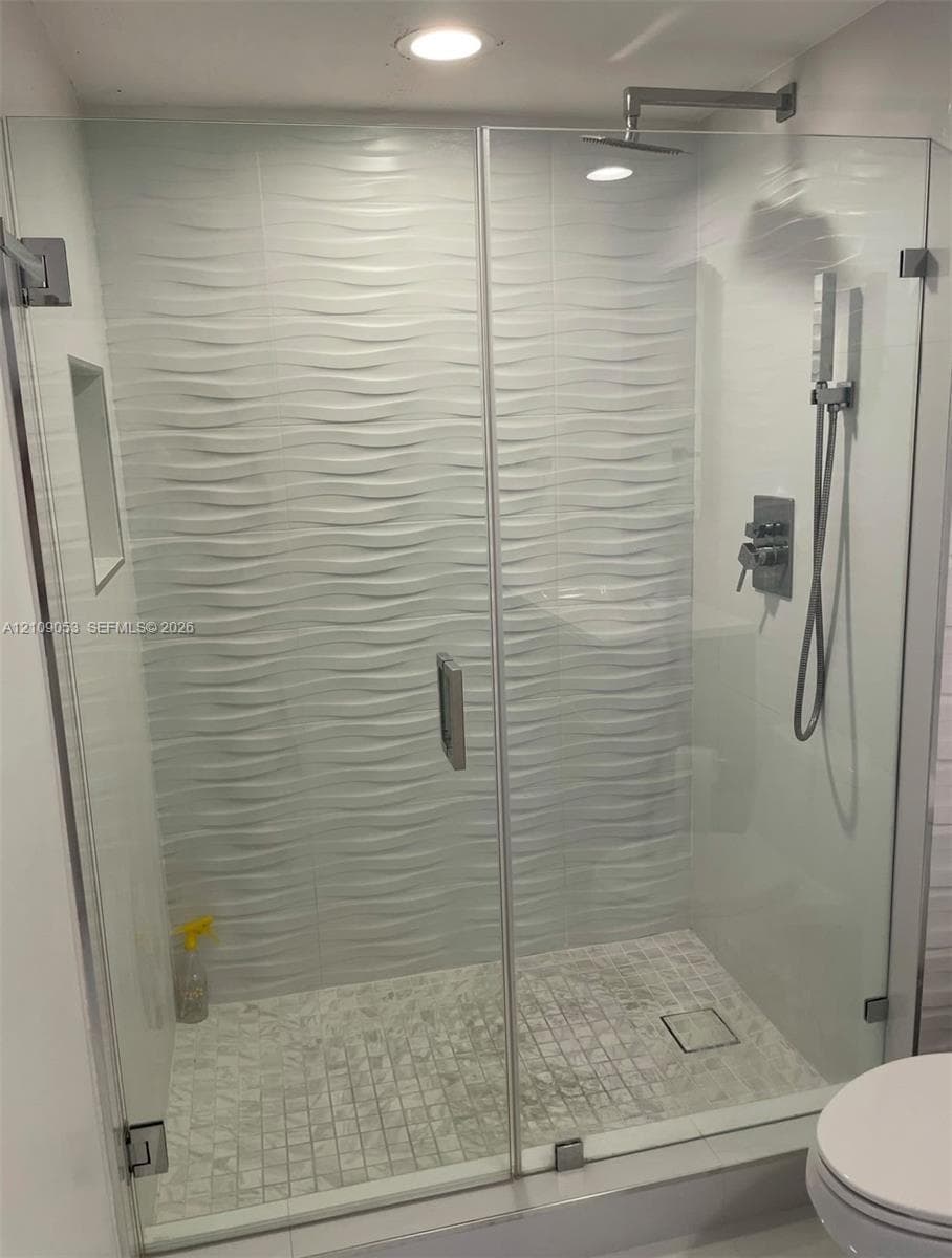 Walk-in shower featuring a glass enclosure, textured wave-patterned wall tile, stone-tiled floor, rainfall showerhead, and a hand-held shower fixture