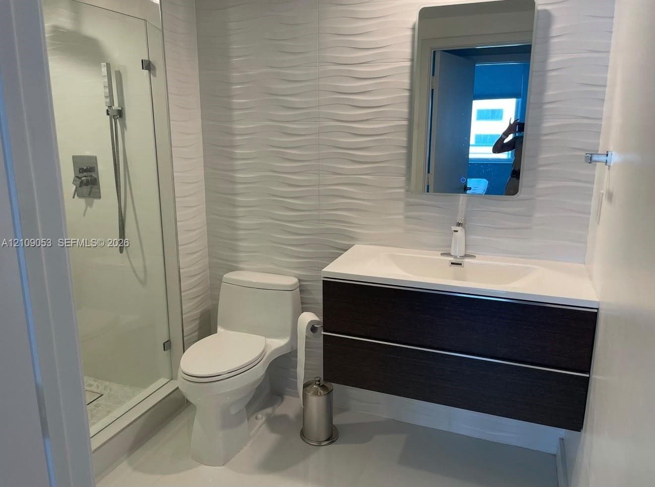 Modern bathroom featuring textured wave-pattern wall tile, a floating wood-finish vanity with an integrated countertop sink, and a glass-enclosed shower with a handheld showerhead