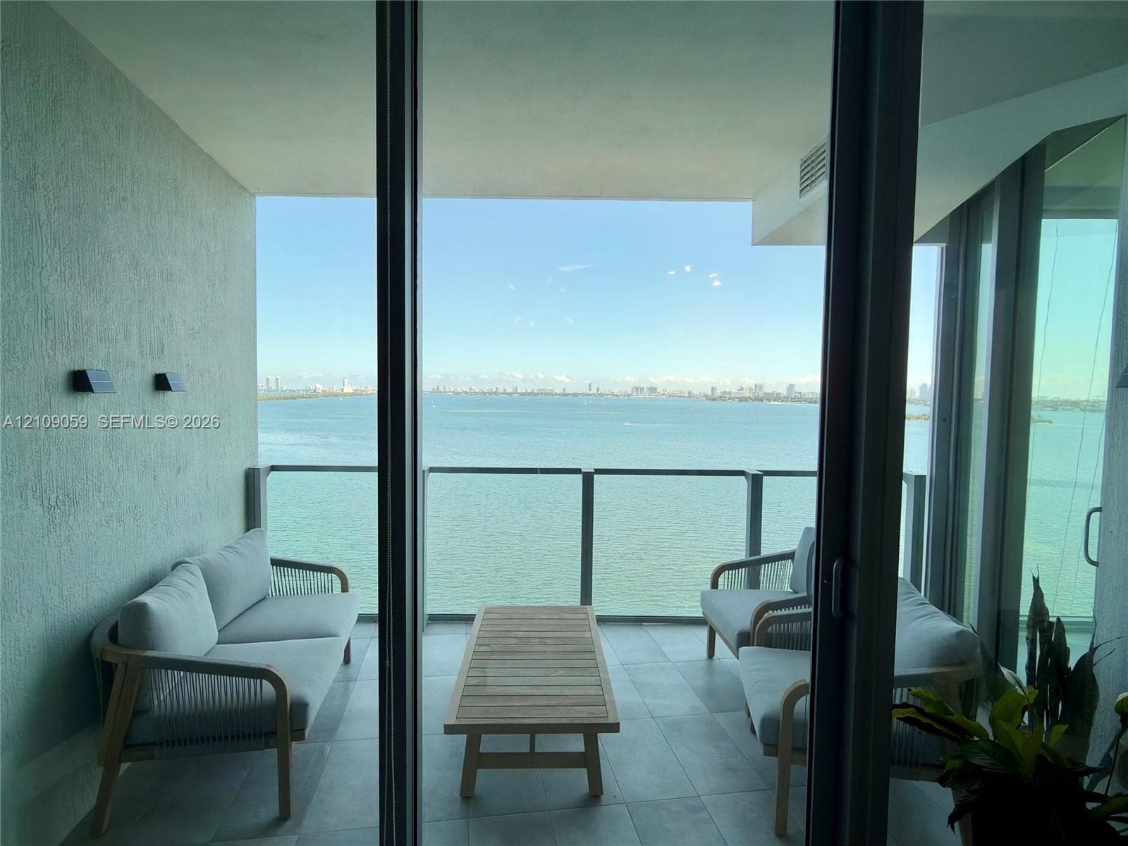 Covered balcony featuring large-format stone-finish floor tiles, wall-mounted exterior lighting, and a glass-paneled railing overlooking the water