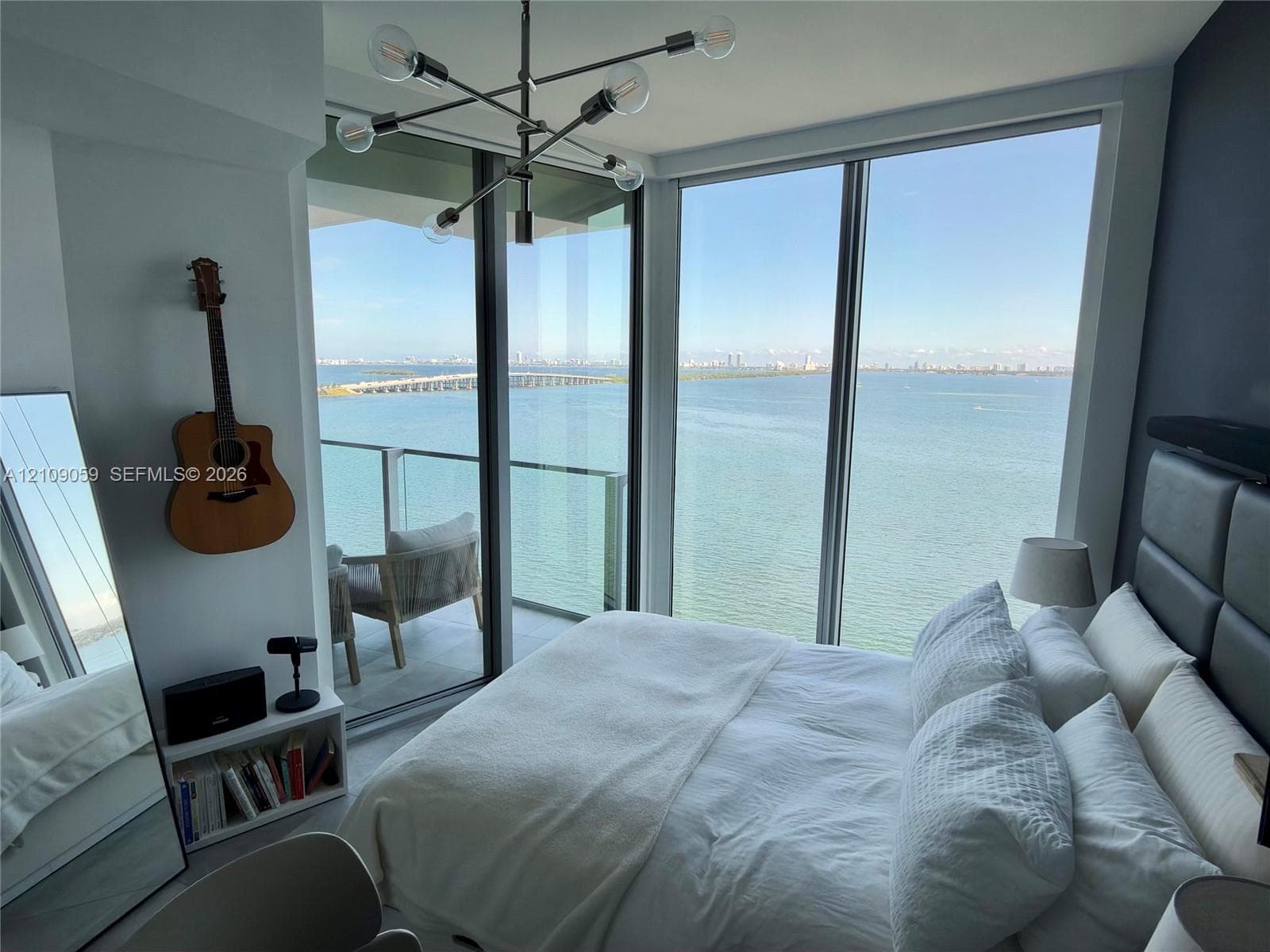 Floor-to-ceiling windows, private balcony with glass railing, contemporary sputnik-style chandelier, and water backdrop