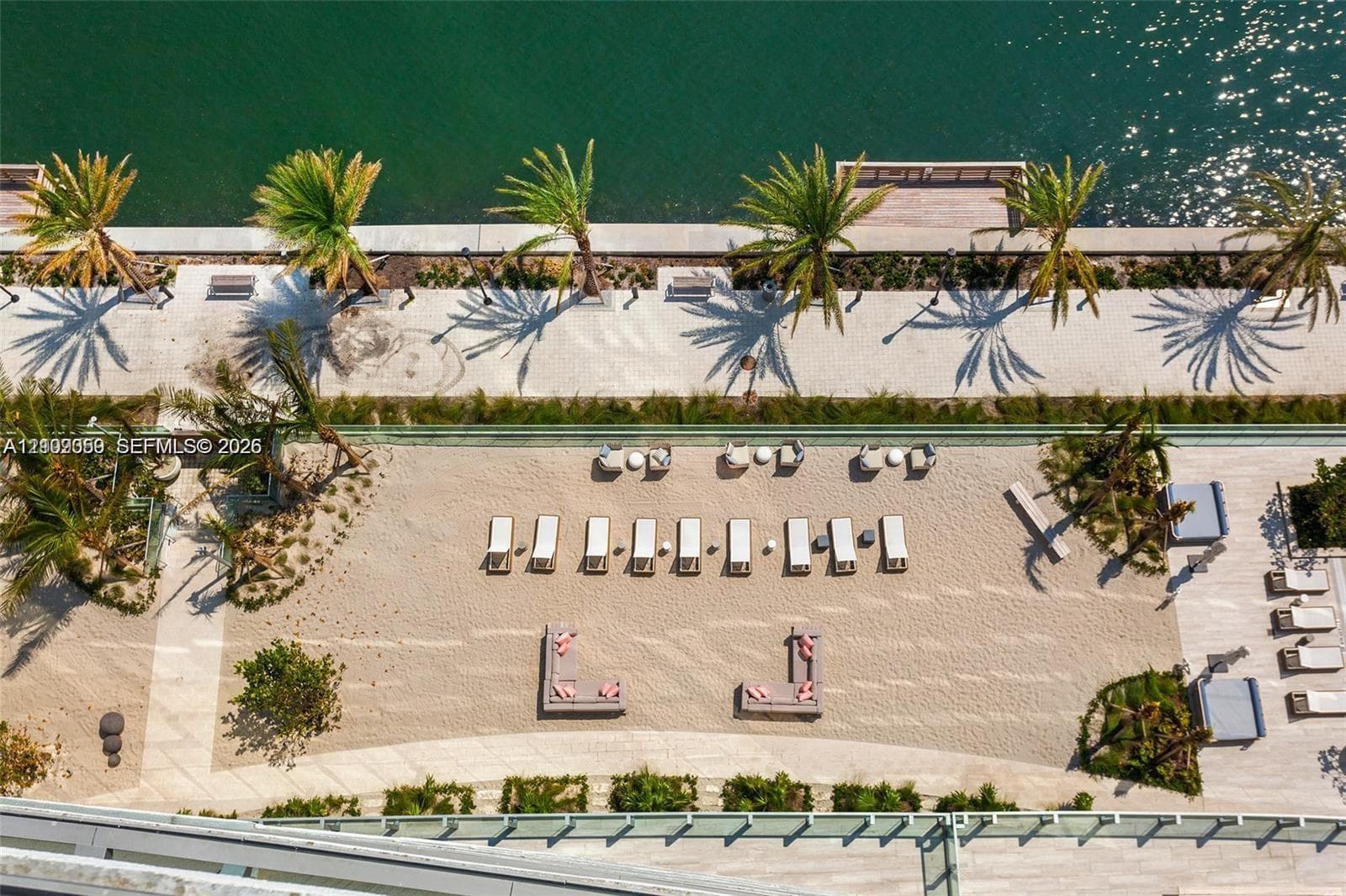 Waterfront recreational area featuring a sandy beach, paved walking path, mature palm trees, and native landscaping