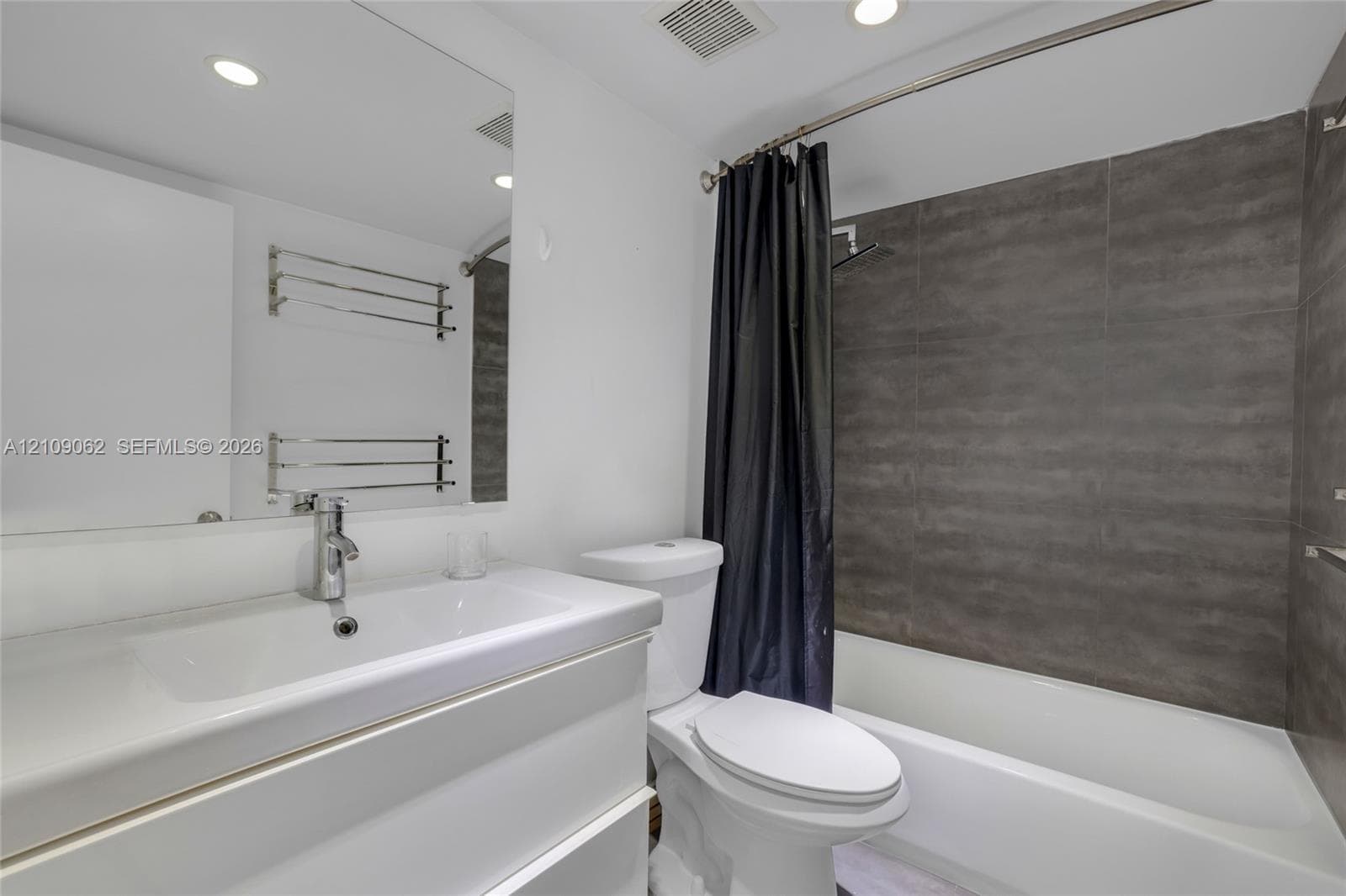Bathroom featuring a white vanity with an integrated sink, polished chrome faucet, large wall-mounted mirror, white commode, and a shower-tub combination with grey tile surround