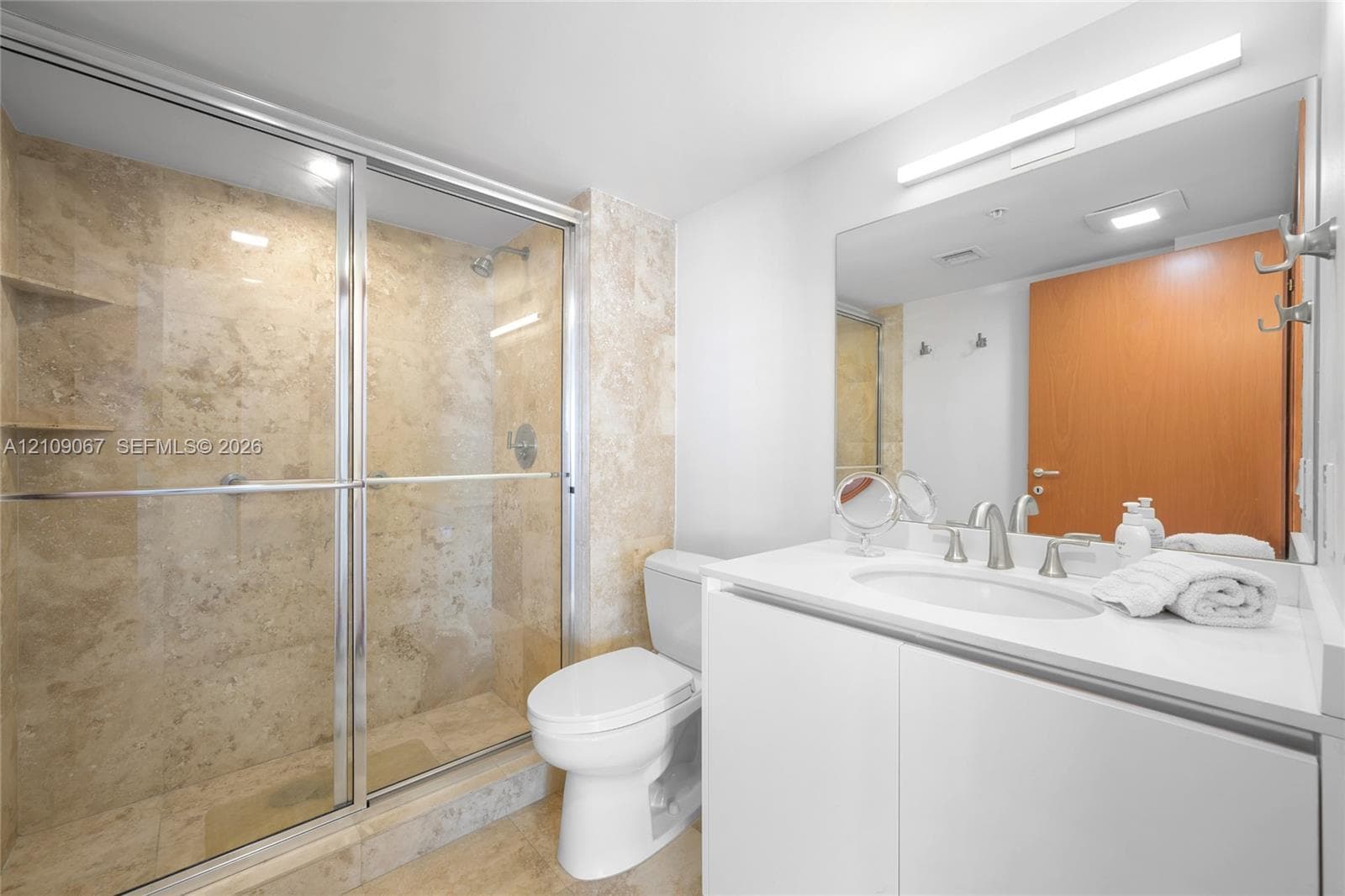 Walk-in shower with stone tile surround, glass sliding door, white vanity with integrated basin, polished metal hardware, and large vanity mirror