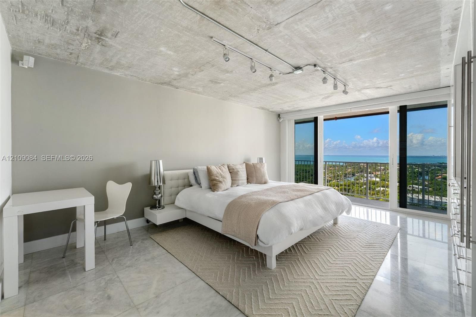 Open-concept living space featuring exposed concrete ceilings, polished stone flooring, custom wall-mounted cabinetry, track lighting, and floor-to-ceiling glass sliding doors with ocean scenery