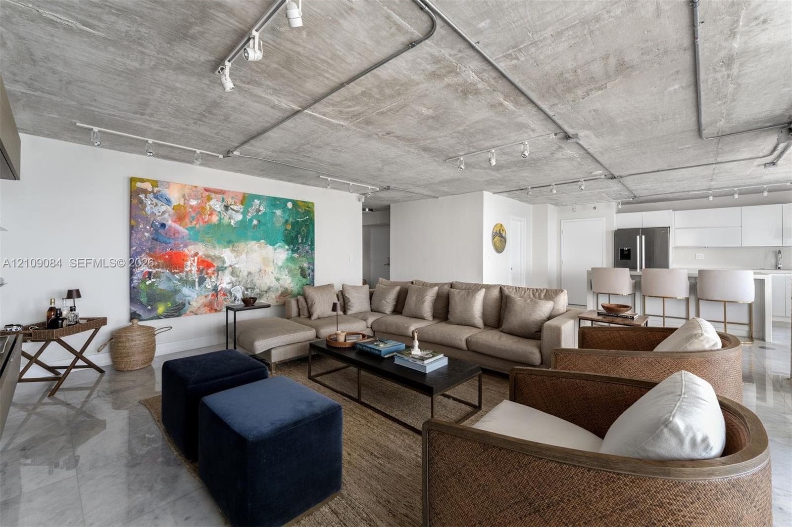 Contemporary kitchen featuring high-gloss white cabinetry, a waterfall-edge quartz island, stainless steel appliances, exposed concrete ceiling with industrial track lighting, and floor-to-ceiling windows with ocean scenery