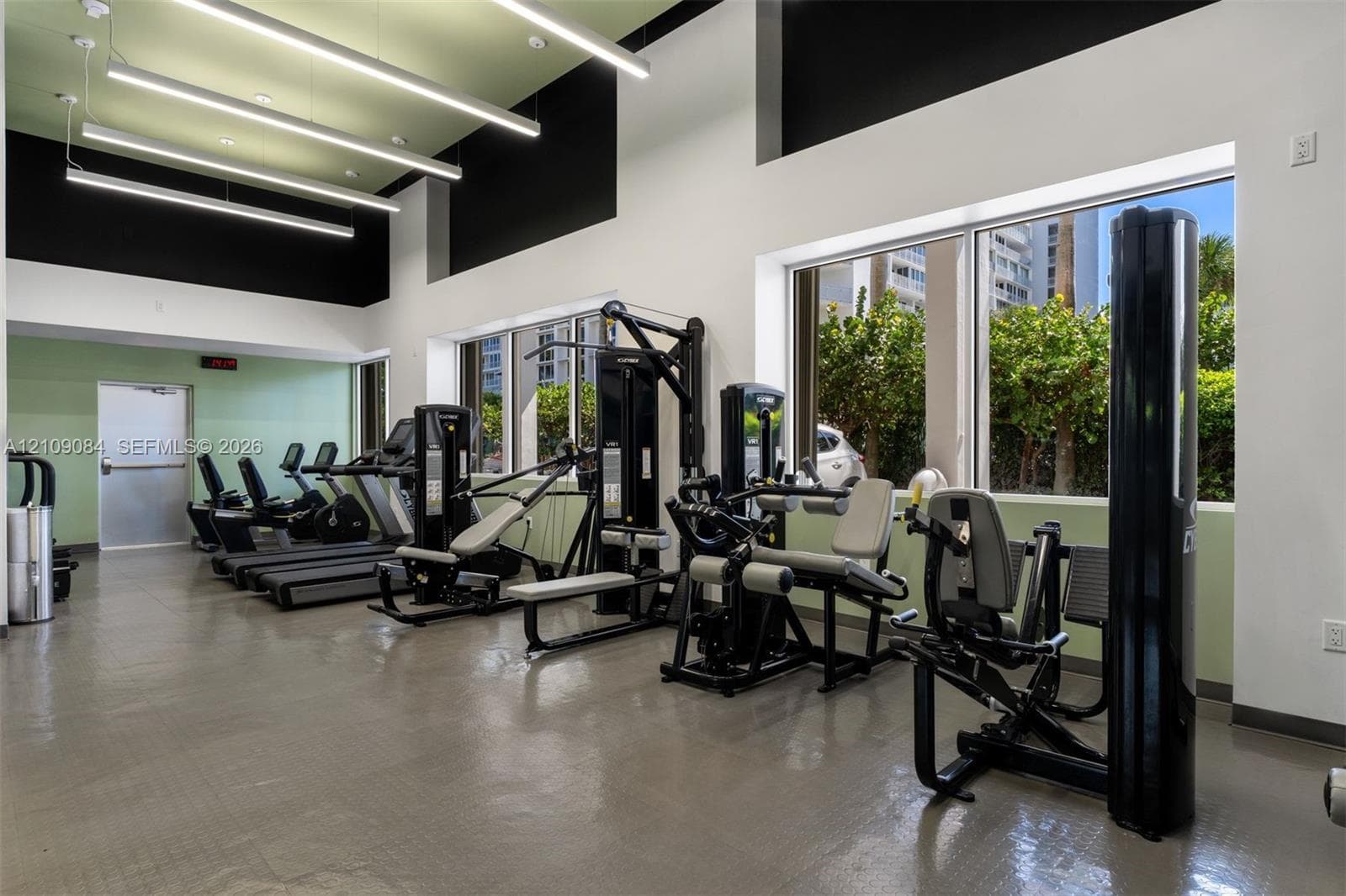 Exposed concrete ceiling with industrial track lighting, floor-to-ceiling glass paneling, built-in high-gloss white cabinetry, polished tile flooring, and panoramic ocean backdrop