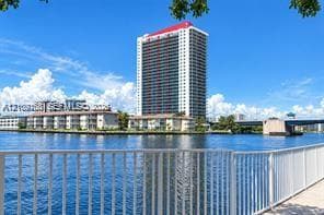 Waterfront high-rise building with a white facade and red roofline, featuring balcony-lined exterior walls and a metal railing along the paved bulkhead