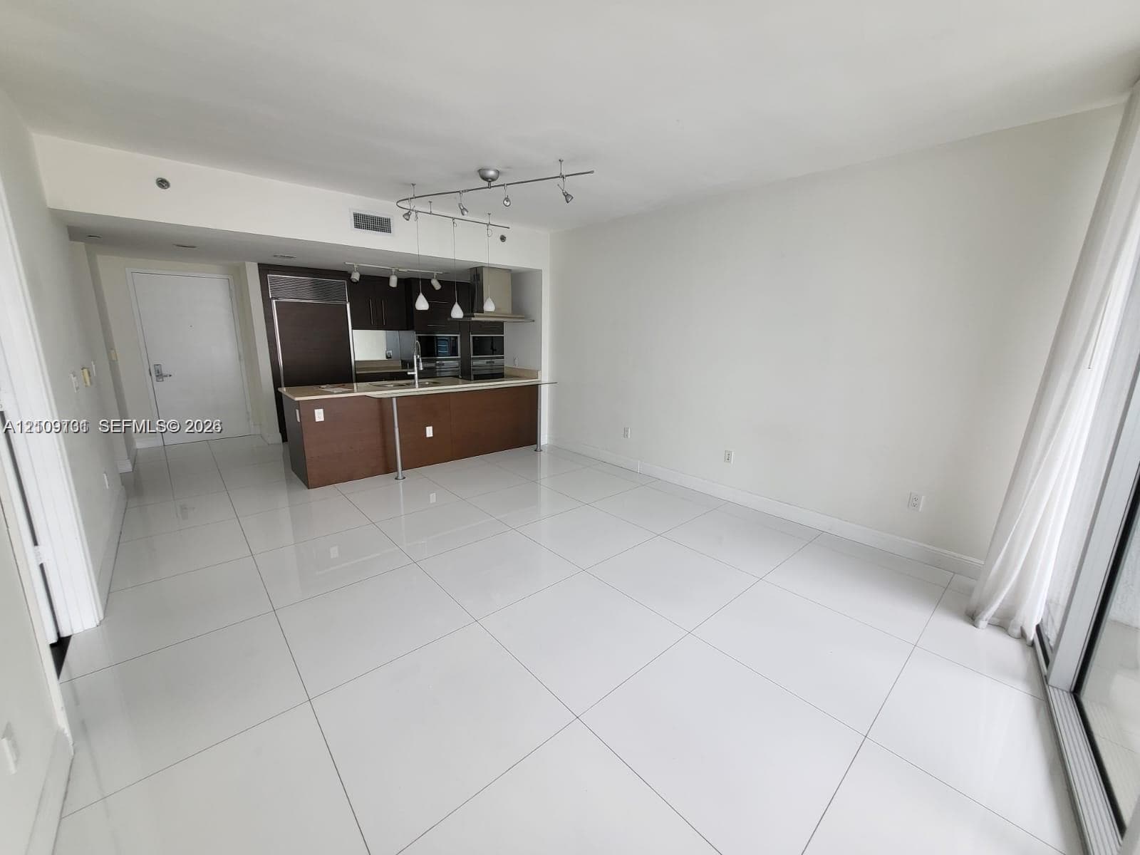 Open-concept kitchen featuring dark wood-finish cabinetry, a breakfast bar with light-toned countertop, stainless steel built-in appliances, track lighting, and white tile flooring