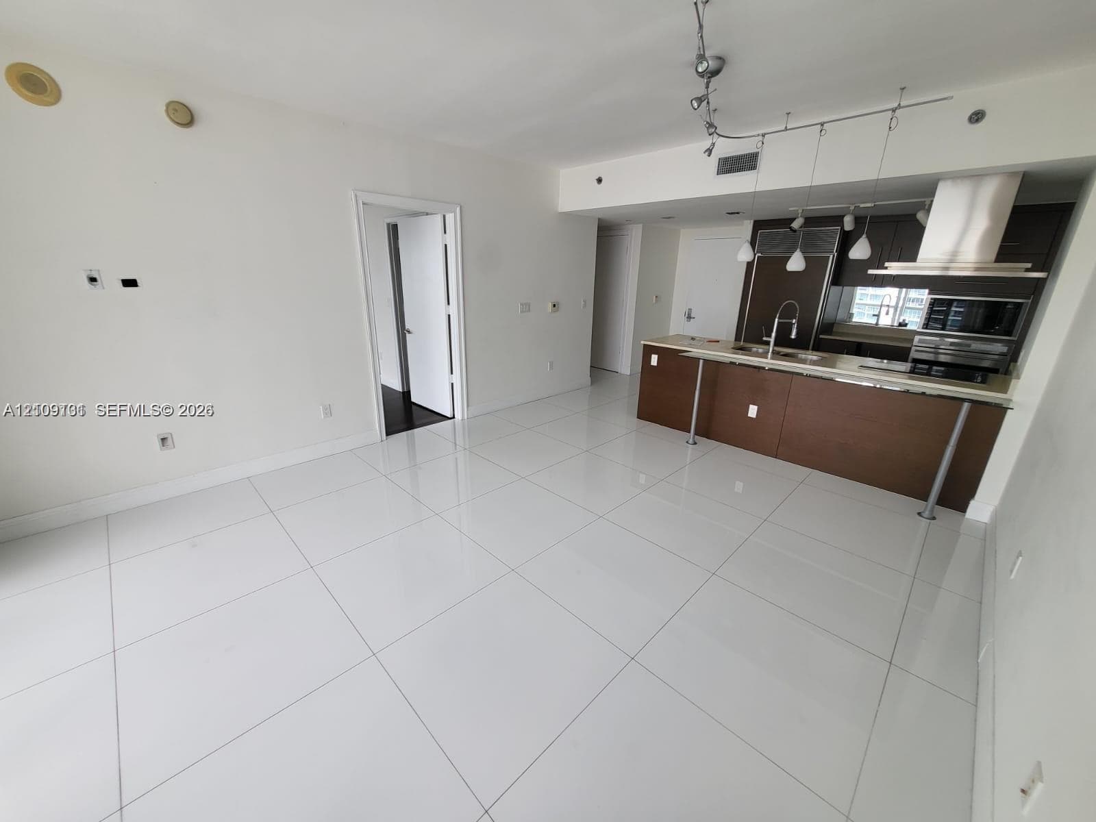 Open-concept living space featuring high-gloss tile flooring, kitchen island with breakfast bar, dark-finish cabinetry, stainless steel built-in appliances, and track lighting