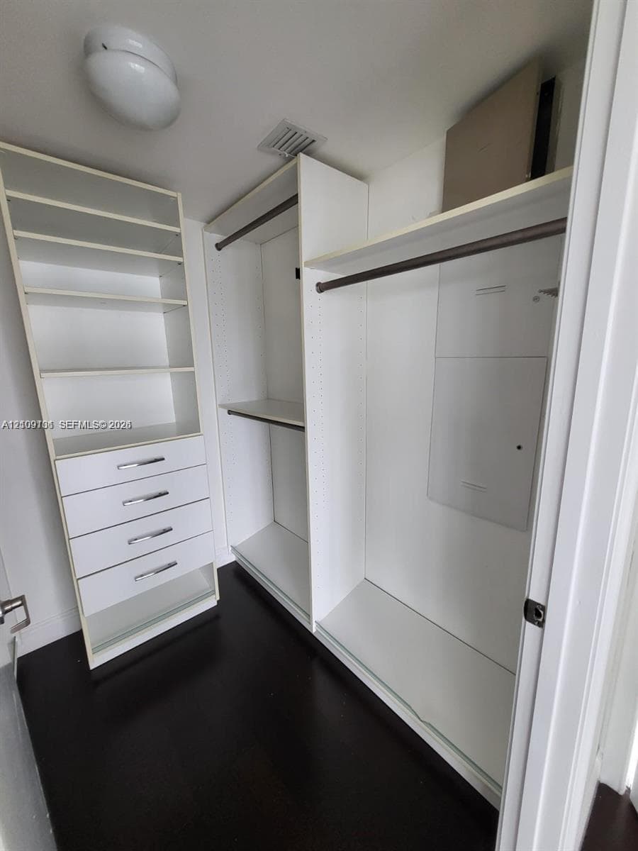 Custom walk-in closet featuring white built-in shelving, four pull-out drawers with silver-tone hardware, two garment rods, and dark-finish flooring