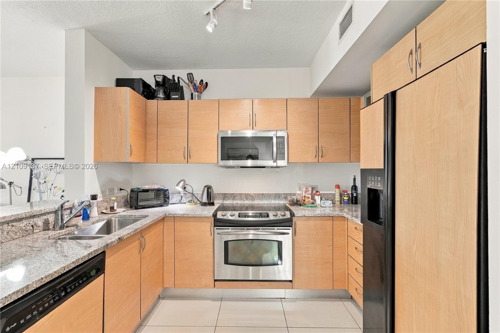 Kitchen featuring light-toned wood cabinetry, stone-surface countertops, stainless steel appliances, a dual-basin stainless steel sink, and tile flooring