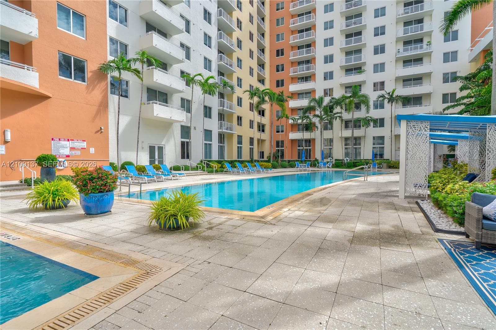 Swimming pool area featuring a stone-paver pool deck, mature palm trees, multi-level exterior balconies, and a covered pergola structure