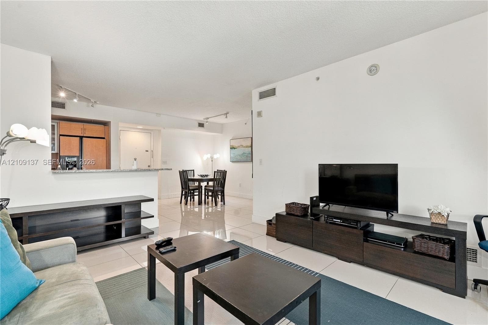 Open-concept interior featuring light-toned tile flooring, recessed track lighting, and a kitchen pass-through counter with stone-finish surfacing