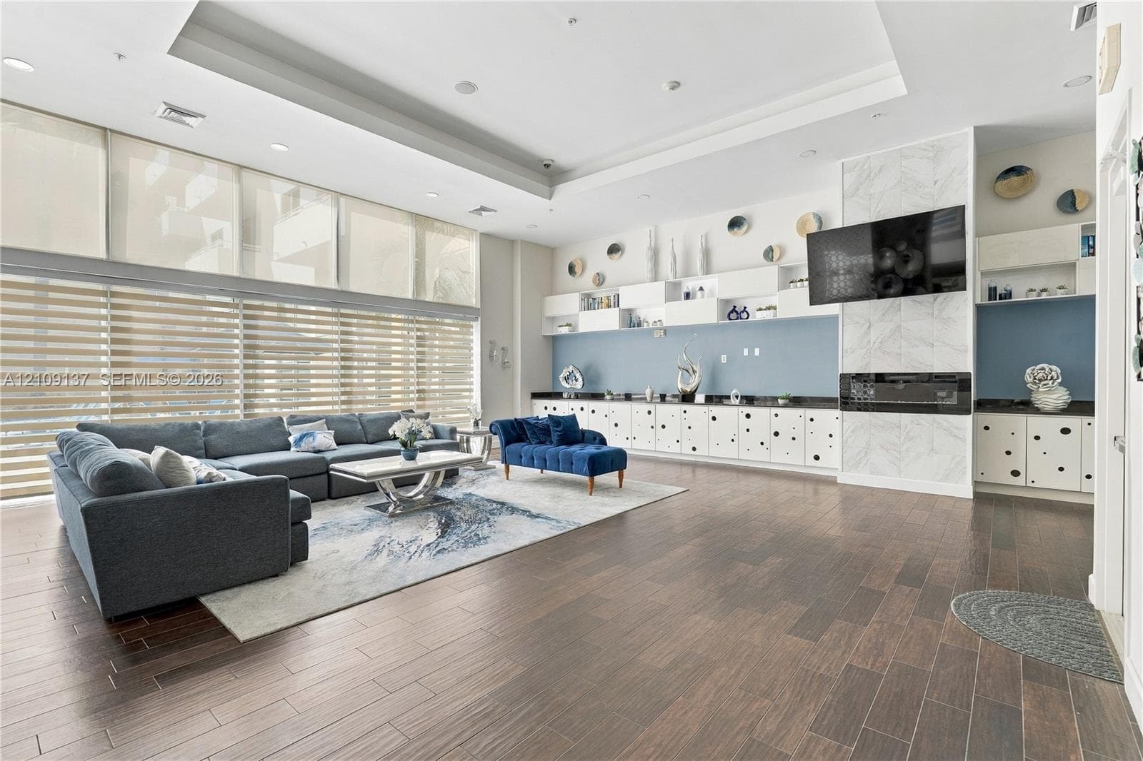 Expansive living space featuring wood-finish flooring, floor-to-ceiling windows with roller shades, recessed tray ceilings, built-in cabinetry, and a fireplace with stone-finish surround