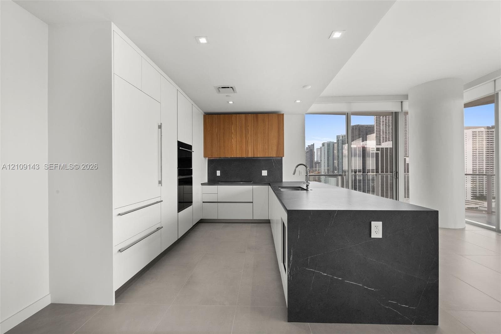 Kitchen featuring flat-panel white cabinetry, wood-finish upper cabinets, expansive waterfall-edge stone island, built-in wall oven, and large-format tile flooring