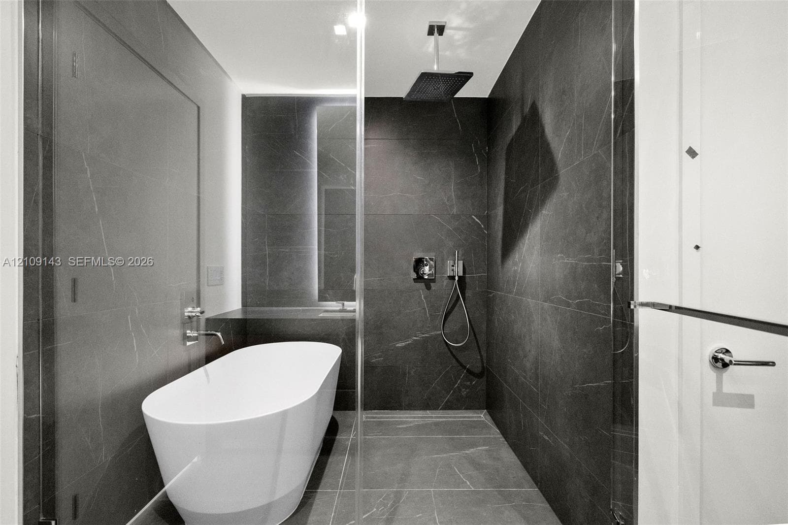 Freestanding soaking tub, large-format dark tile wall surfacing, overhead rainfall showerhead, handheld shower fixture, and frameless glass enclosure