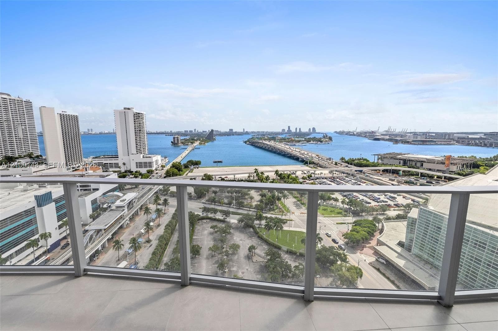 Outdoor balcony with neutral tile flooring and a glass-paneled railing overlooking a cityscape and waterway