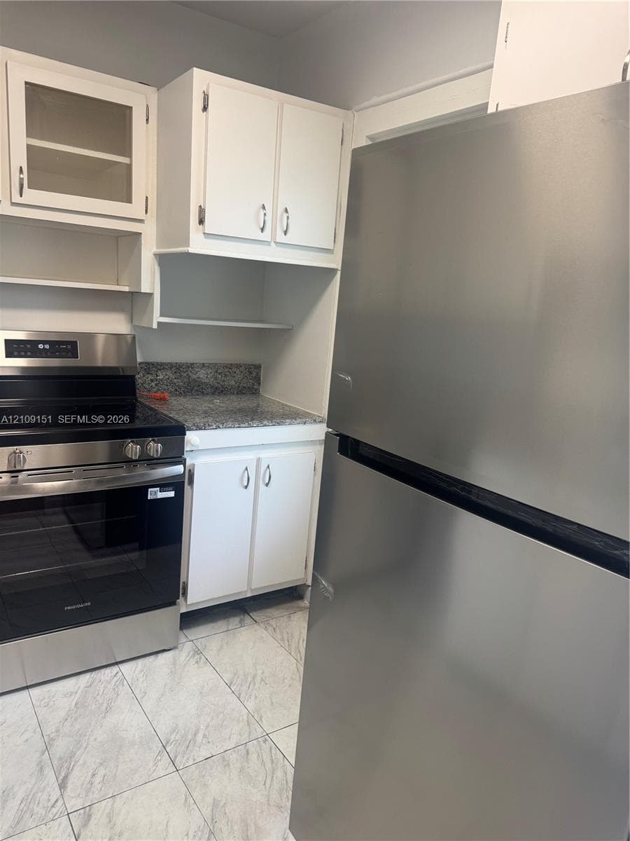 Kitchen featuring white shaker-style cabinetry with glass-front display panels, stone-finish countertops, stainless steel appliance suite, and light-toned tile flooring