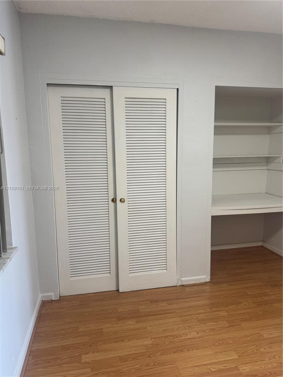 Closet featuring white louvered bifold doors with metallic hardware and integrated recessed shelving units with a built-in desktop surface over wood-finish flooring