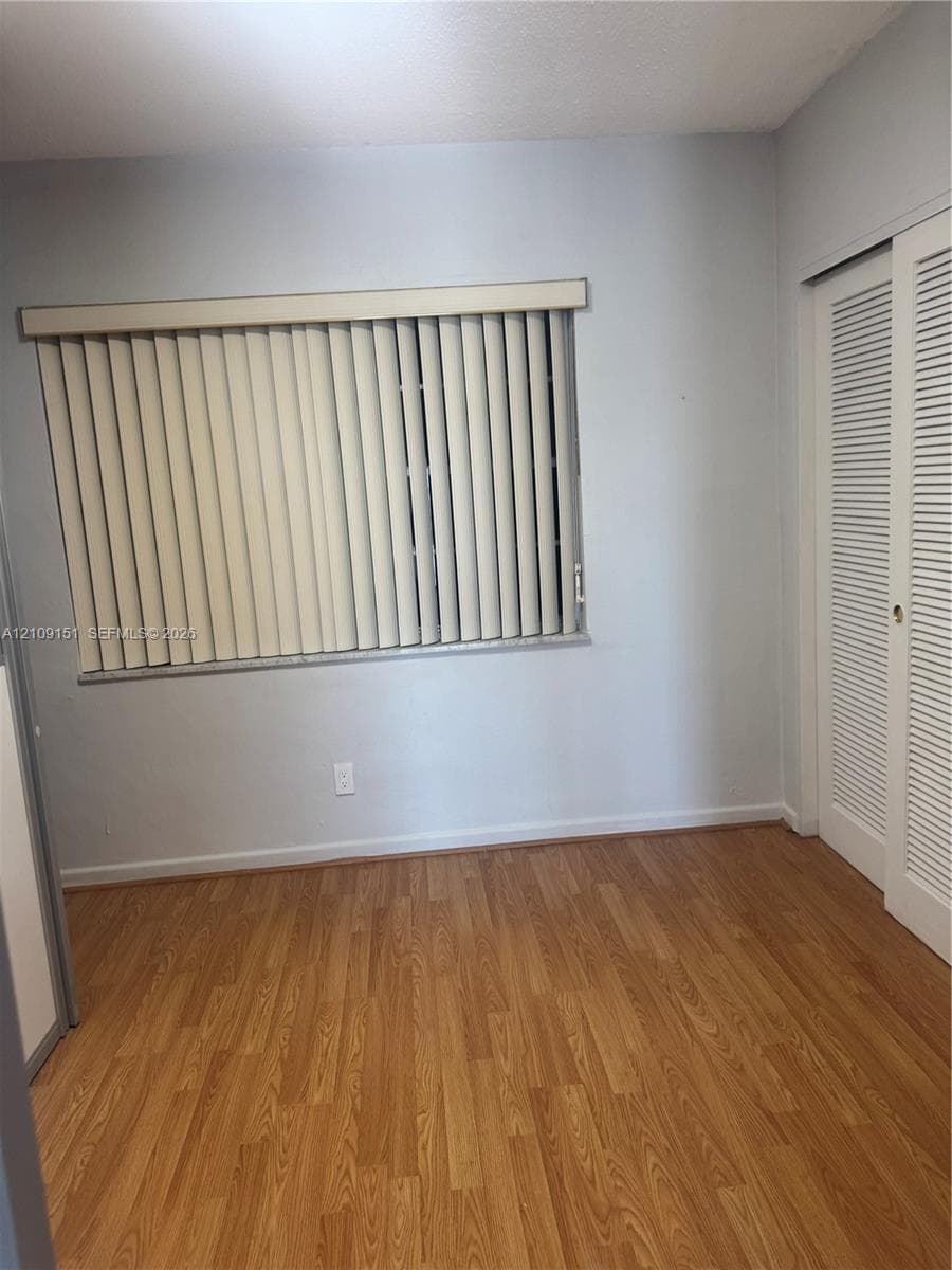 Room featuring wood-finish flooring, neutral wall paint, a window with vertical blinds, and a closet with louvered double doors