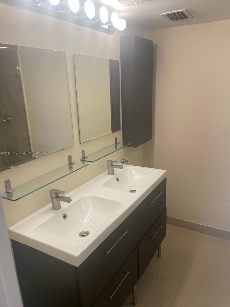 Dual-sink vanity with deep-tone cabinetry, metallic hardware, rectangular drop-in basins, glass shelving, and wall-mounted vanity lighting