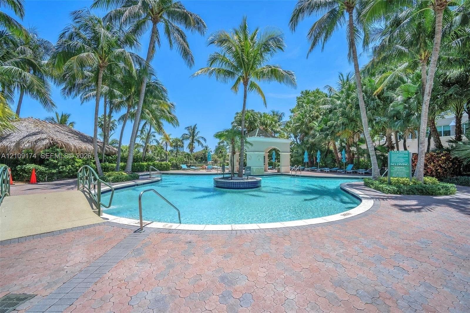 Swimming pool featuring a central island, paver deck, pedestrian bridge, and gazebo structure, surrounded by mature palm trees