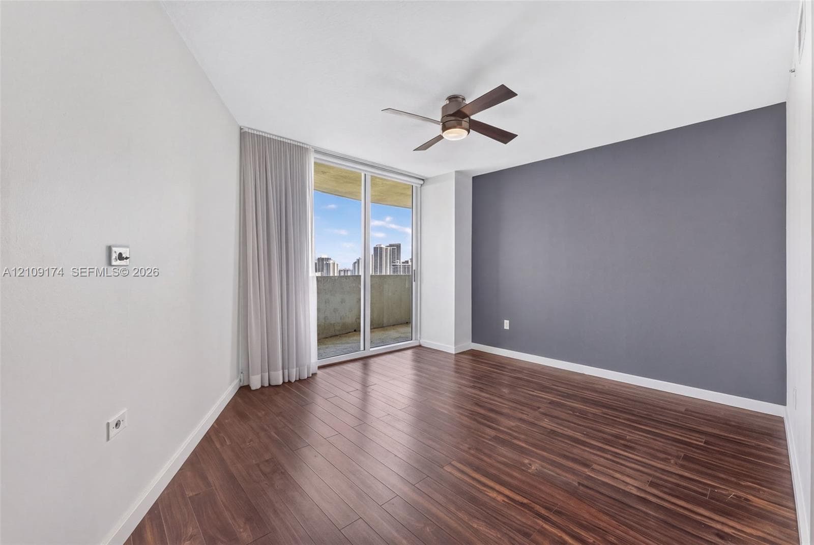 Wood-finish flooring, dark grey accent wall, ceiling fan with integrated light fixture, and floor-to-ceiling sliding glass doors opening to a balcony