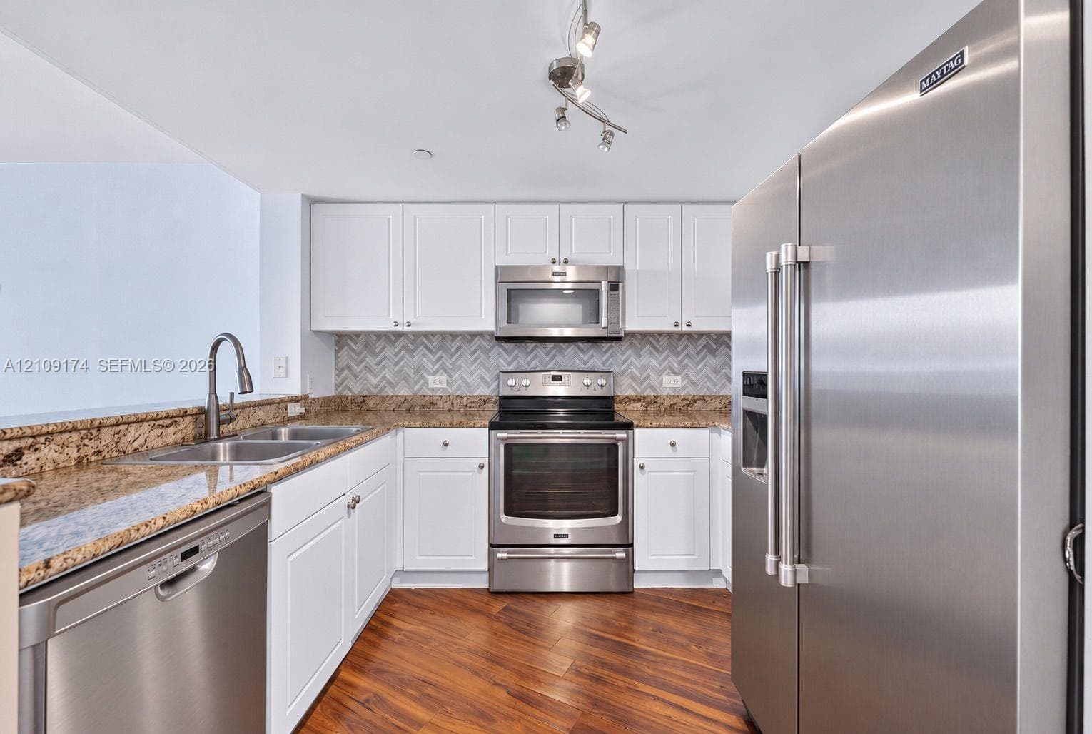 Kitchen featuring white shaker-style cabinetry, stone countertops, stainless steel appliances, a herringbone-patterned backsplash, and wood-finish flooring