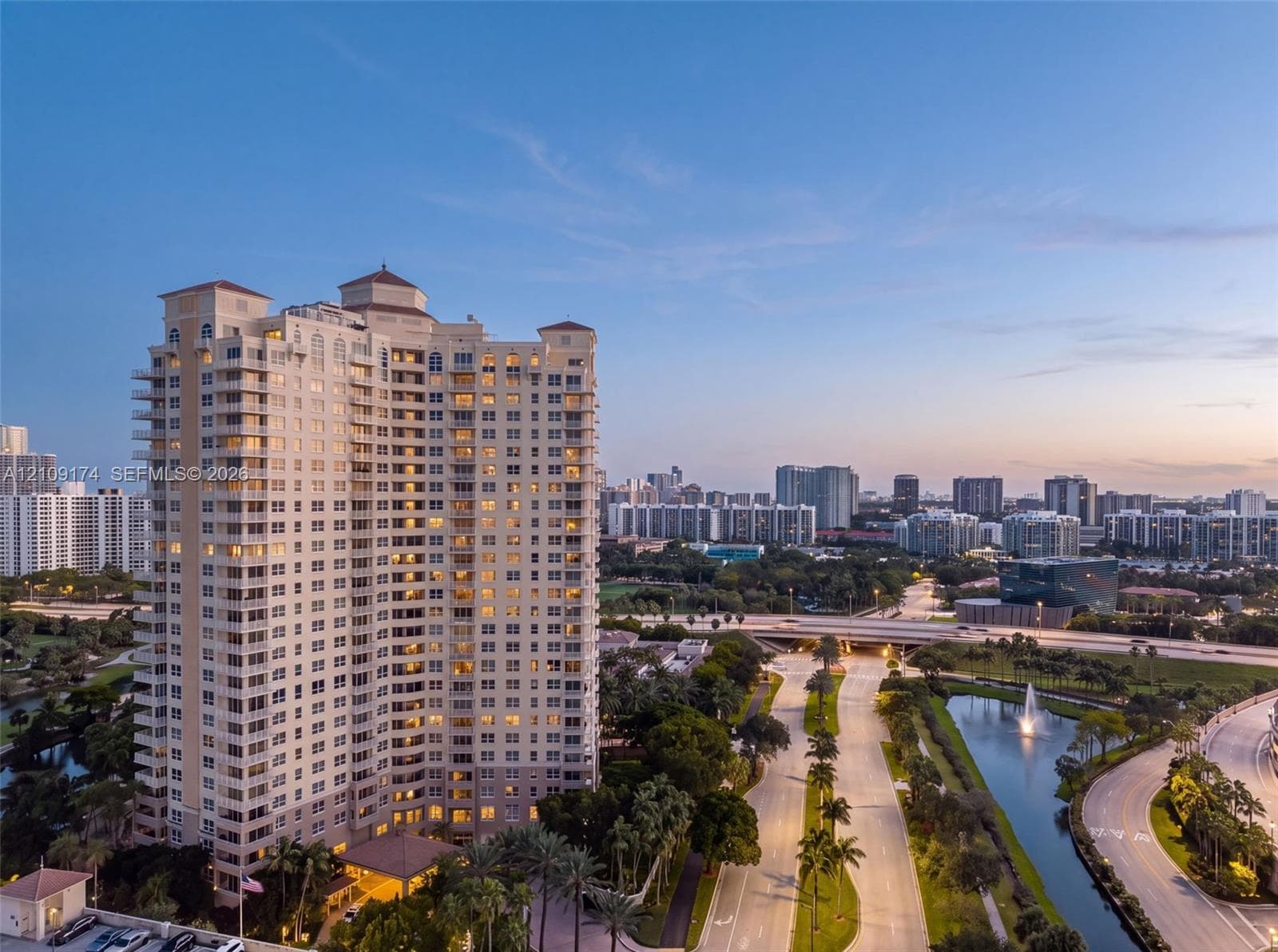High-rise residential tower featuring a tiered architectural design, private balconies, a covered porte-cochère entry, mature palm tree landscaping, and a surrounding water feature
