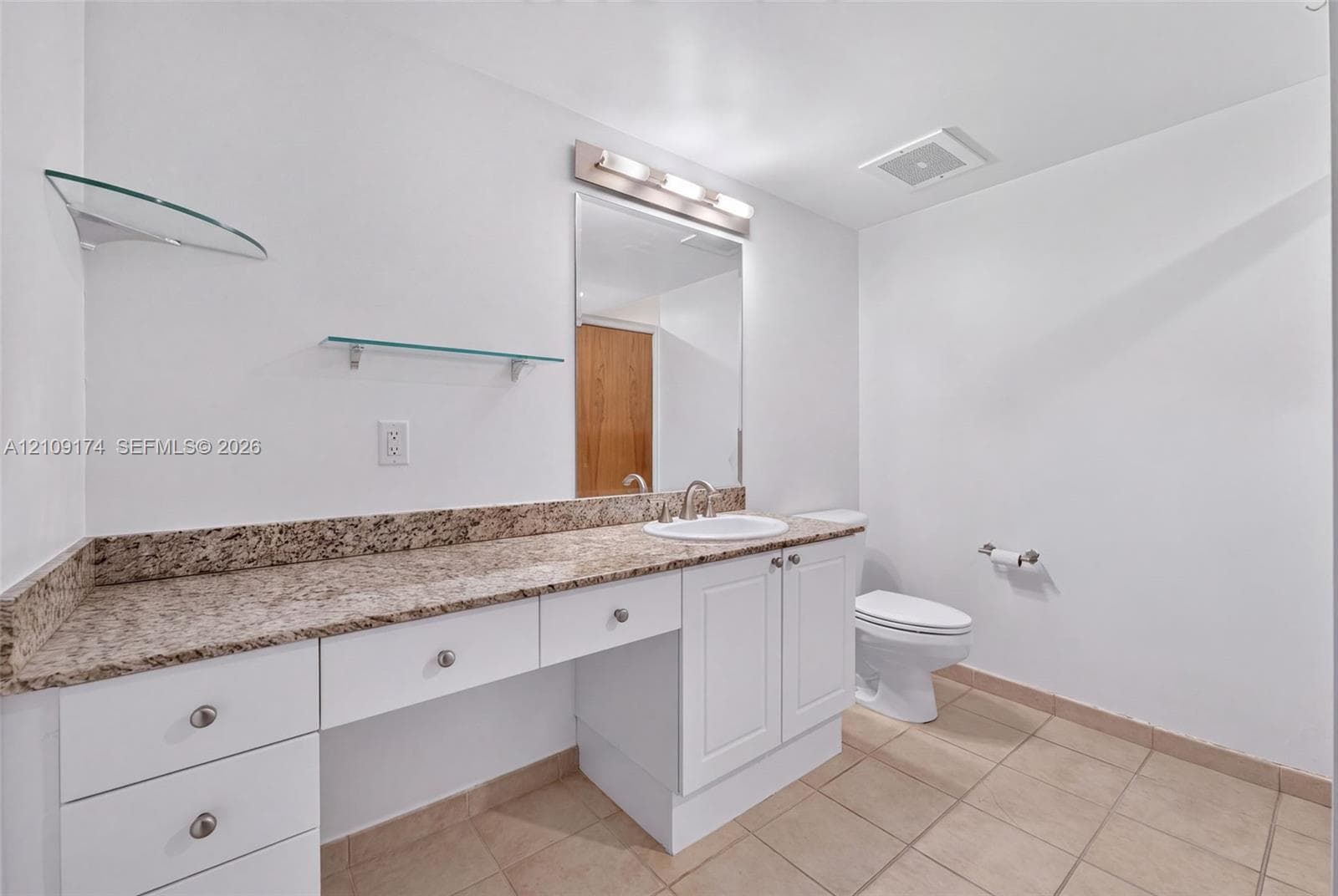 White cabinetry with stone-finish countertop, undermount sink, integrated vanity lighting, glass shelving, and ceramic tile flooring
