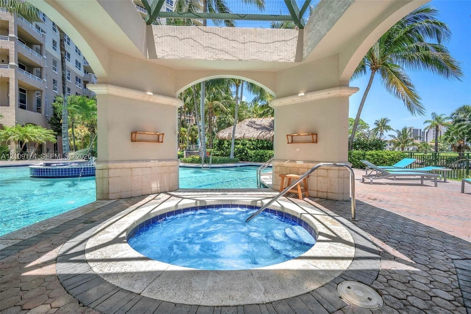 Covered spa area featuring stone paver flooring, architectural pillars with stone accents, built-in wall-mounted lighting, and a metal safety handrail
