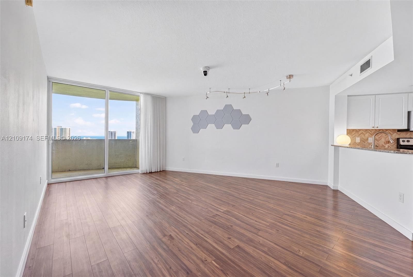 Open-concept living space featuring wood-finish flooring, floor-to-ceiling sliding glass doors, a private balcony, track lighting, and a kitchen pass-through with stone-finish countertops